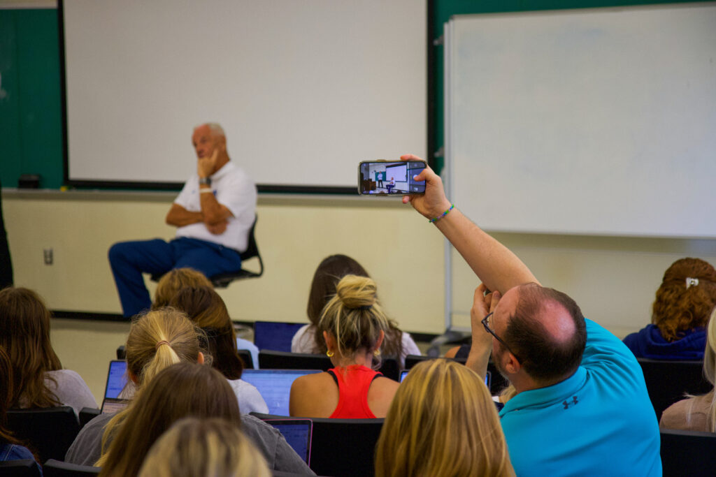 a man, sitting in a full classroom, takes a photo of Roy Williams on his phone