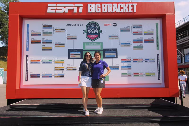 two women stand in front of a large-scale bracket for a baseball tournament