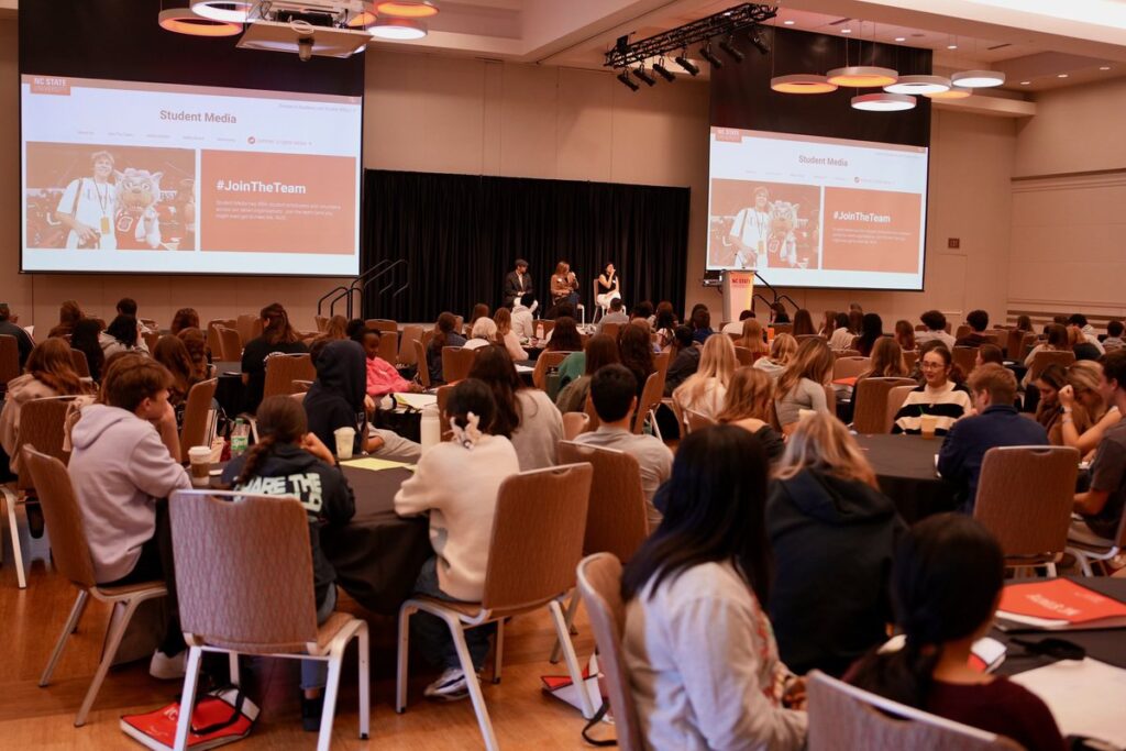 three people sit on a stage in a conference room full of round tables with students. 