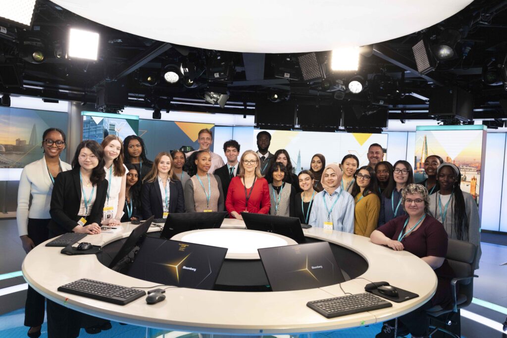 a group of students and program leaders stand behind a news desk in the news studio