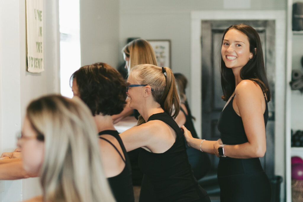 a woman looks at the camera while she supports the back of another woman who is doing a Pilates move