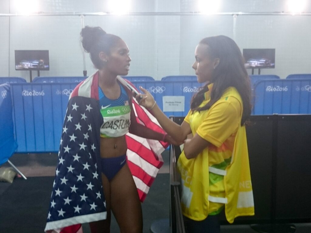 a woman interviews a female track runner wearing an American flag over her shoulders