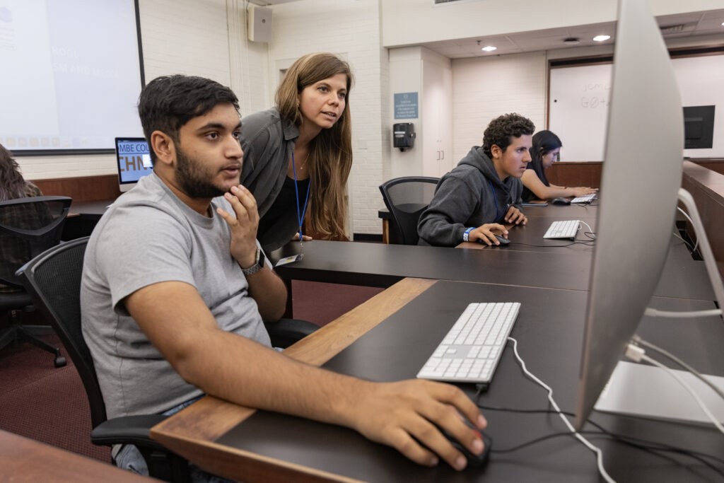Two people look at a computer screen as two people in the background work on their own computers in a classroom in Carroll Hall.