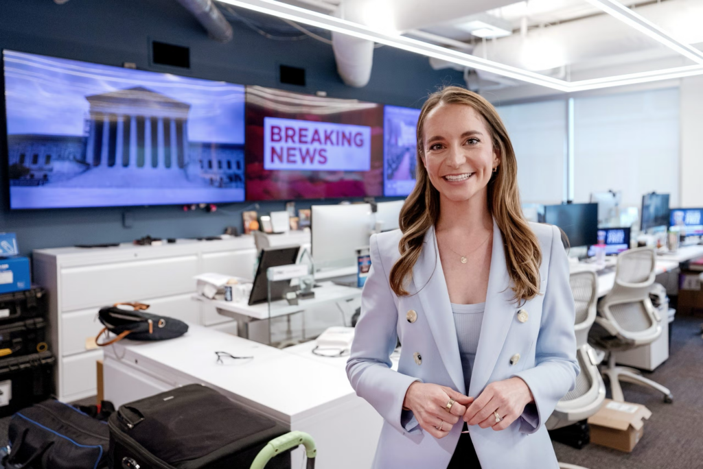 Megan Cassella stands in front of desks and breaking news sign.