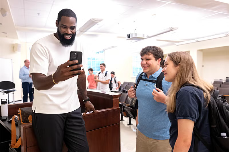 Antawn Jamison holds a phone as he stands with two students in a classroom.
